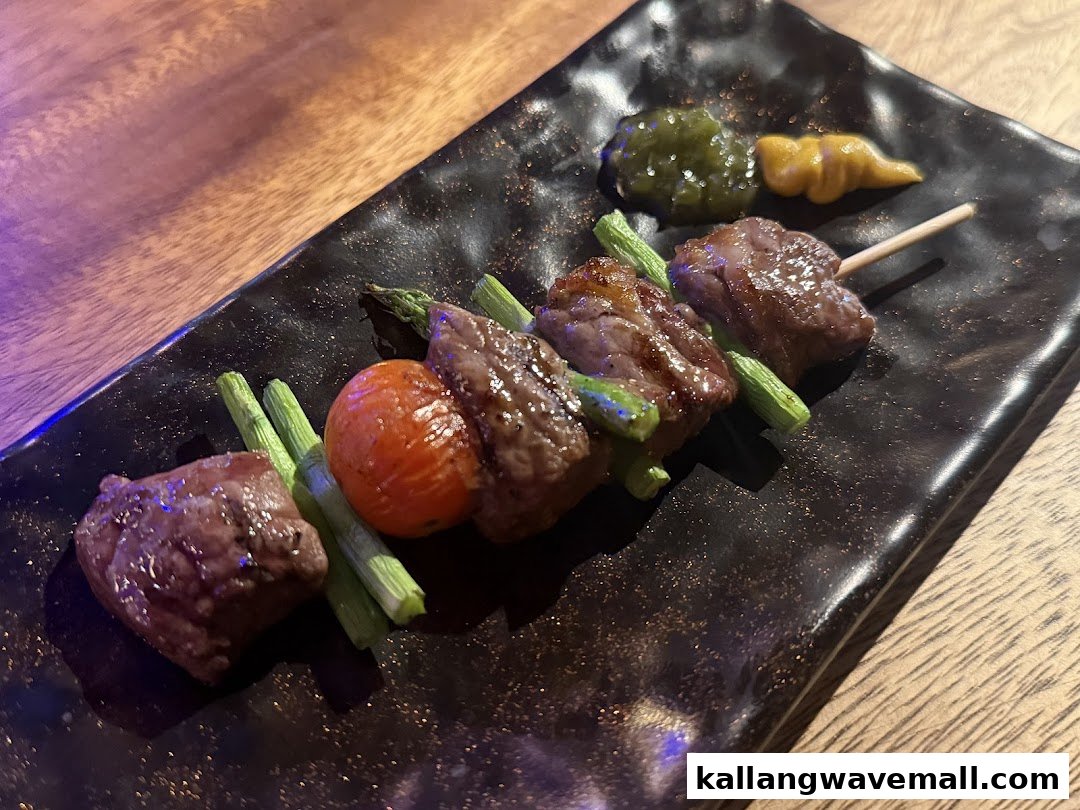 A grilled meat skewer with asparagus and tomato, served with two sauces on a dark plate.
