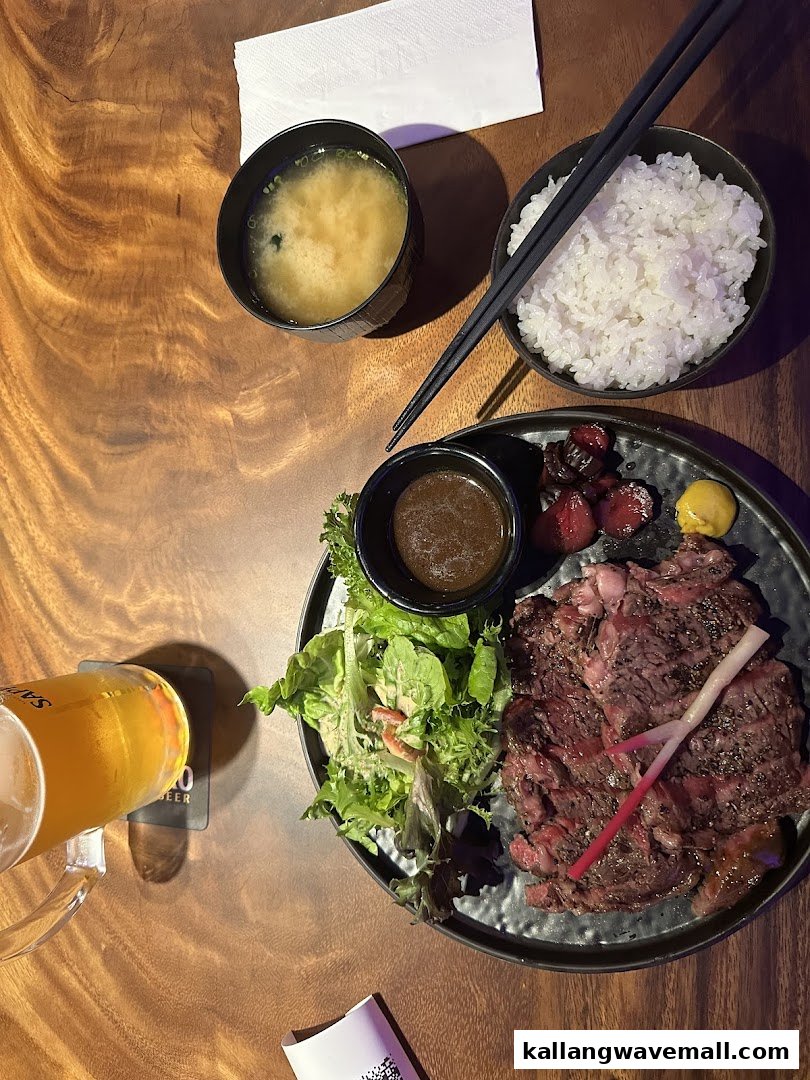 A delicious meal featuring grilled steak, fresh salad, rice, miso soup, and a beer on a wooden table.