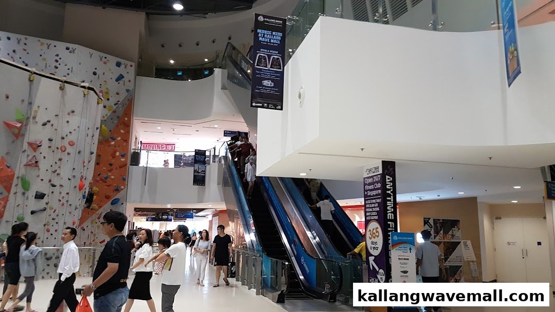 An indoor climbing gym with a large rock wall, escalators, and people in a bustling mall environment.