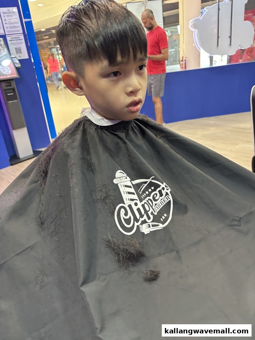 A young boy with a fresh haircut, wearing a Clippers Barber cape.