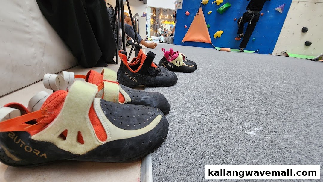 Climbing shoes rest on the floor of a bouldering gym, with a climber visible on the wall in the background.