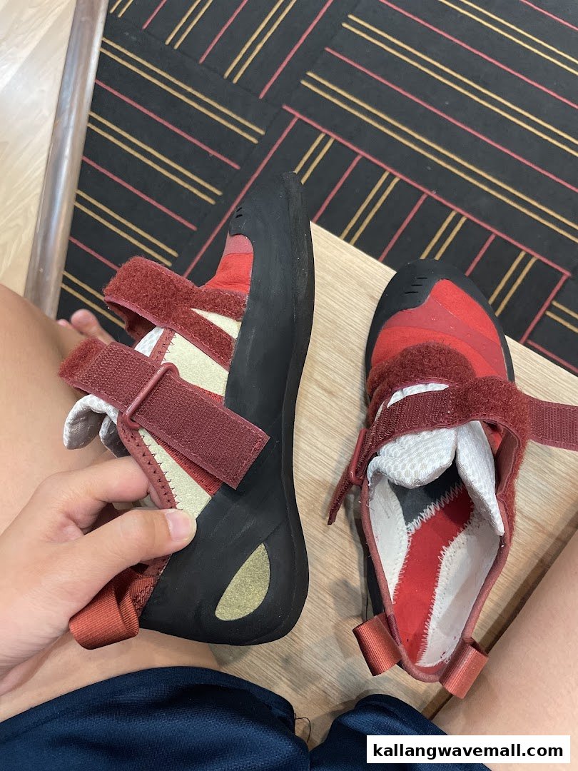 A pair of red and black climbing shoes, one held by a hand, on a wooden surface.