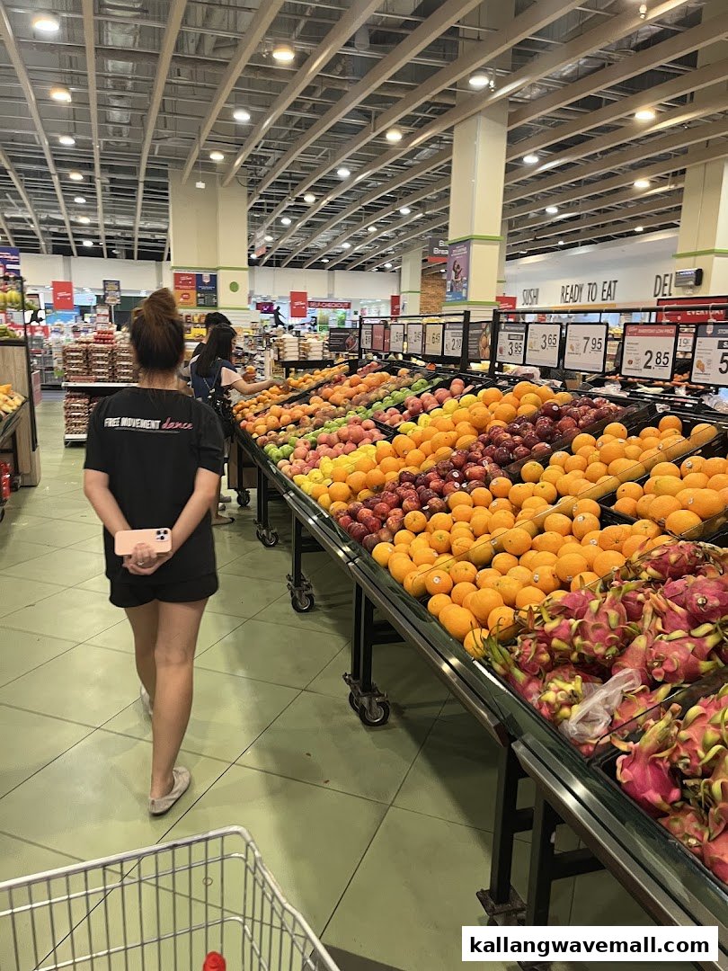 A shopper walks past vibrant displays of fresh fruits, including oranges, apples, and dragon fruit, in a well-lit supermarket aisle.