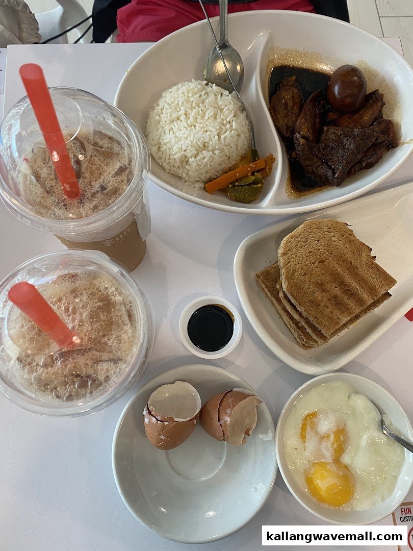 A table spread featuring traditional Singaporean breakfast items like soft-boiled eggs, toast, iced drinks, and a braised meat rice dish.
