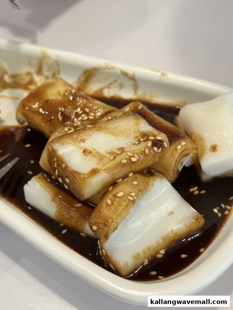 Close-up of rice noodle rolls (Chee Cheong Fun) with sweet sauce and sesame seeds on a white plate.