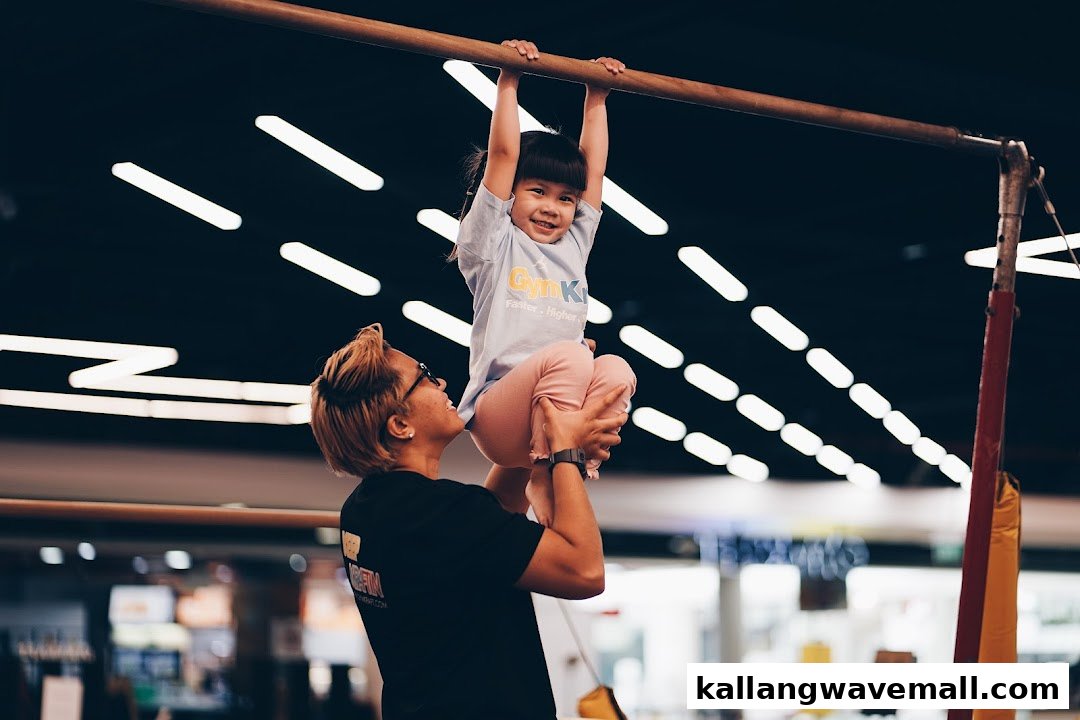 A child smiles while hanging from a gymnastics bar, assisted by an instructor at GymKraft.