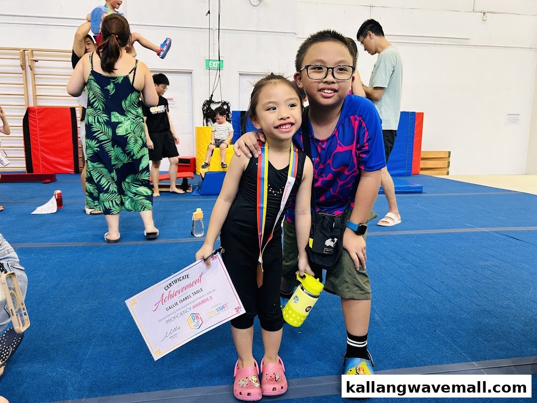 Two children smiling, one holding a gymnastics achievement certificate, inside a vibrant gym facility.