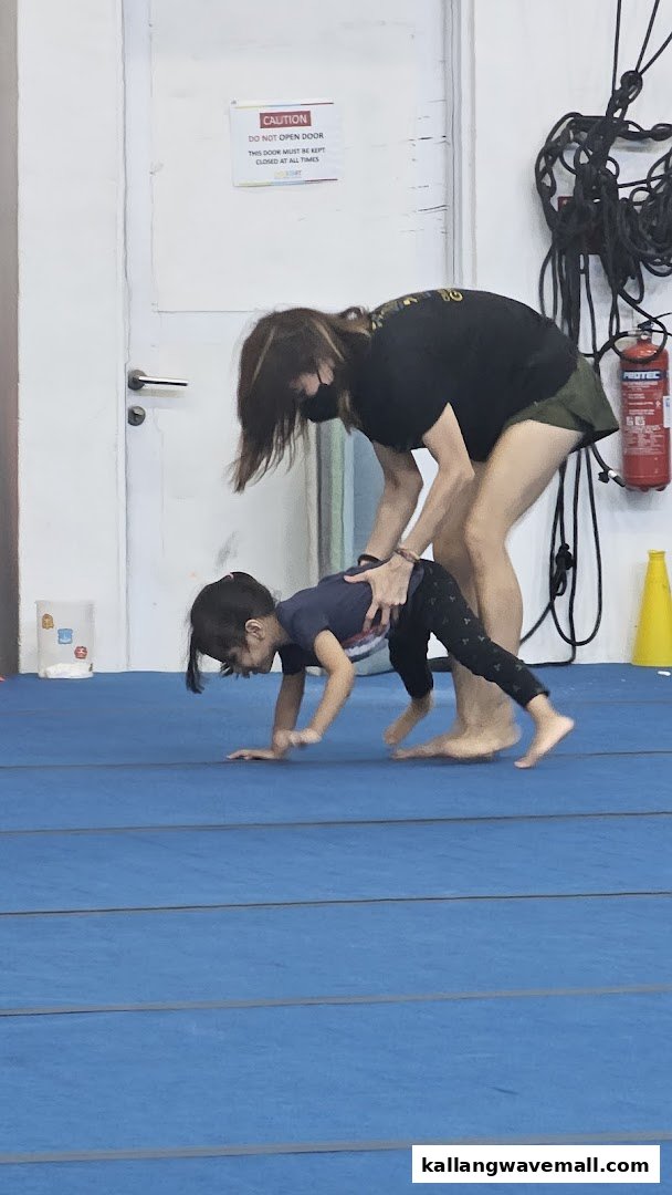An adult assists a child practicing movements on a blue mat in a gym setting.