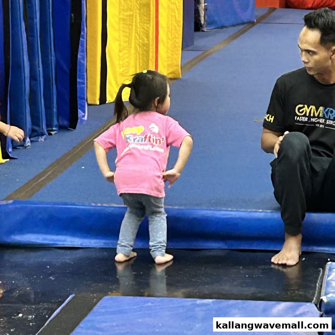 A young child in a pink shirt stands on a blue mat, observed by an adult in a GymKraft shirt.