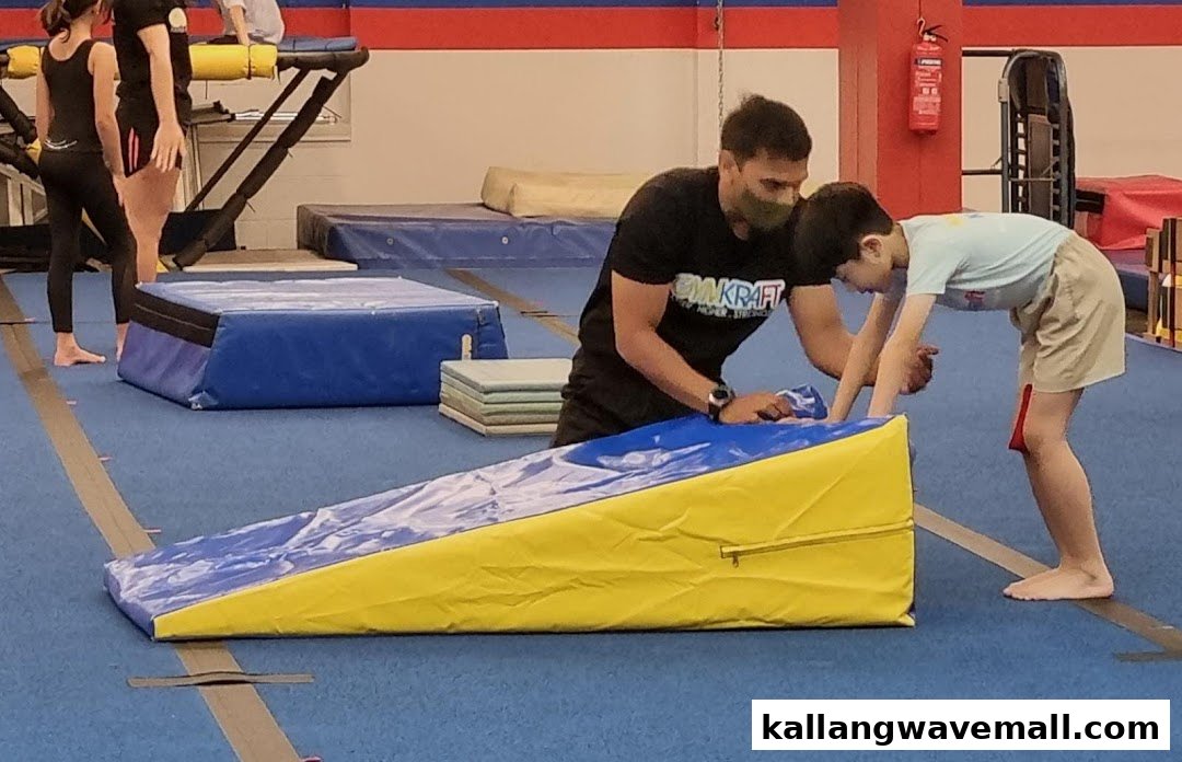 A coach assists a child on a blue mat in a vibrant gym, with other participants in the background.