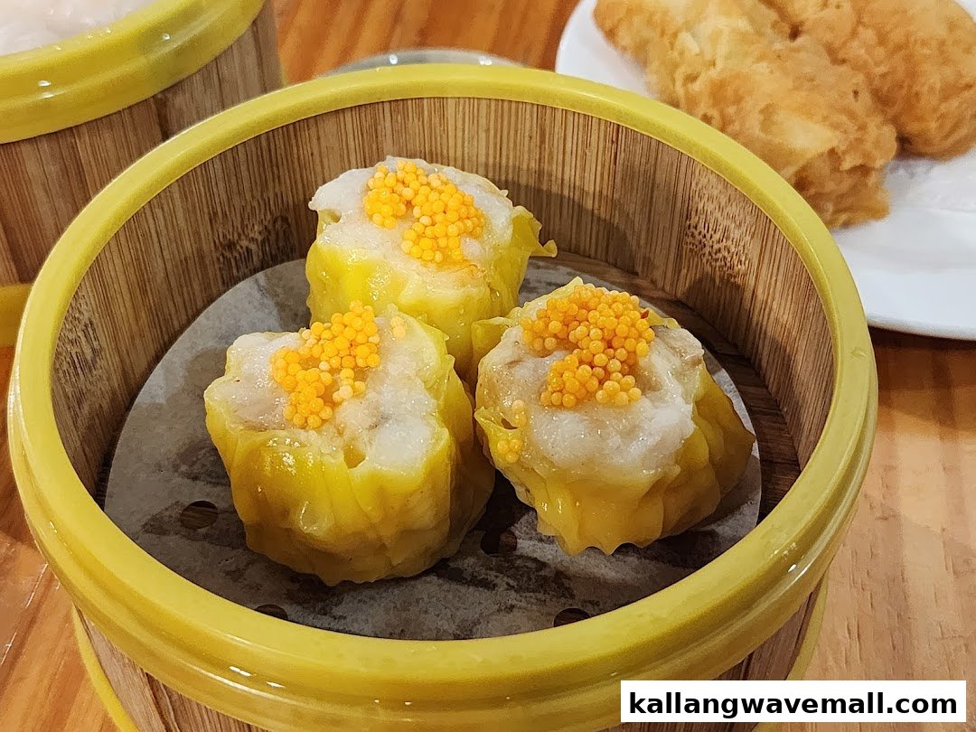 Three vibrant yellow dim sum dumplings, likely siumai, topped with orange roe in a bamboo steamer.