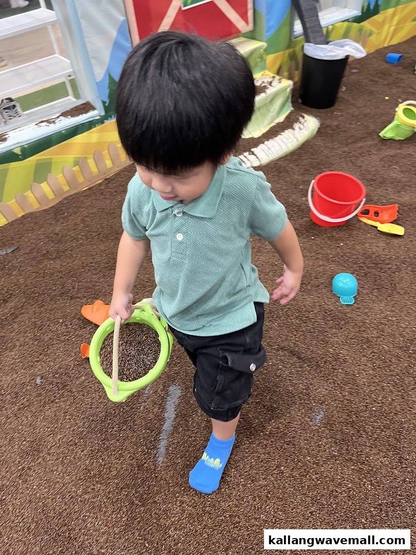 A child plays with a bucket and sieve in an indoor farm-themed play area.