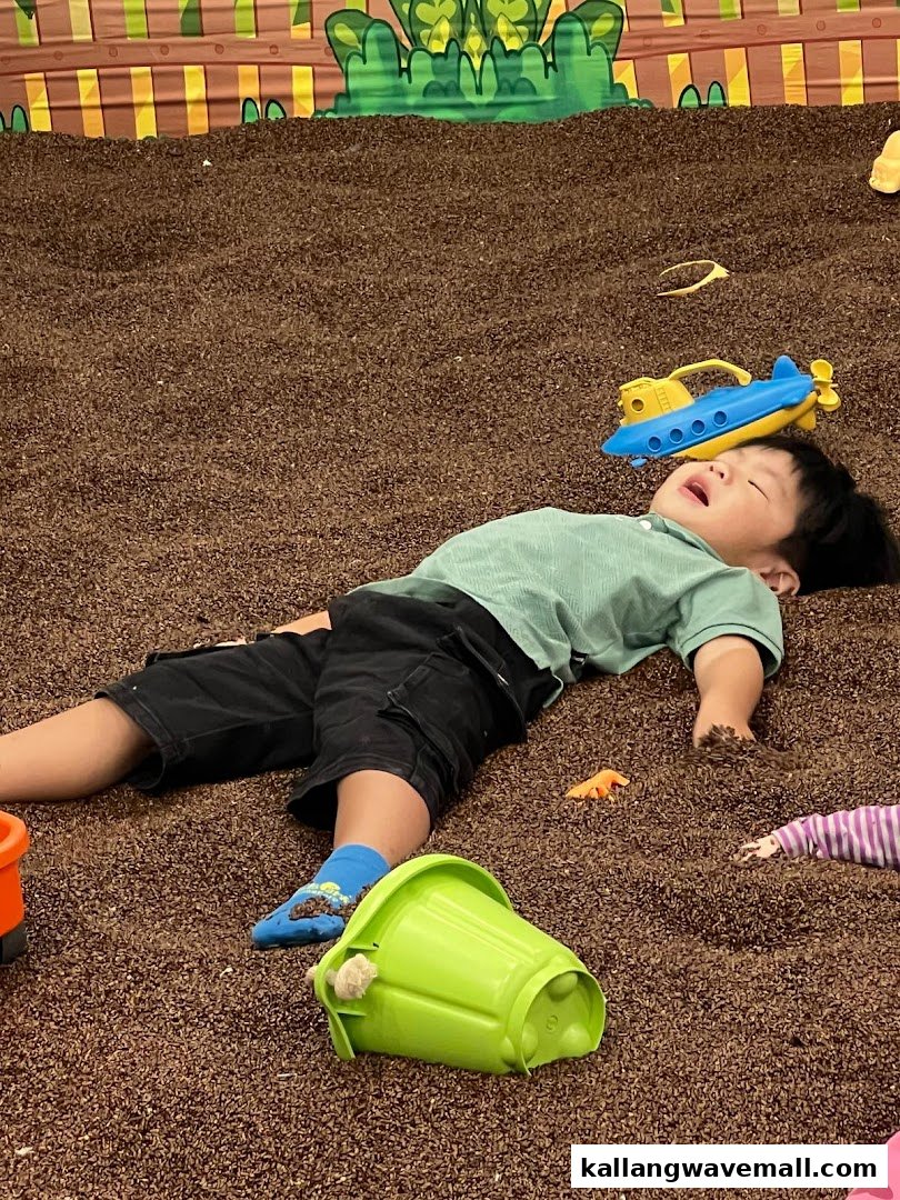 A child rests amidst brown play pellets and toys in a vibrant indoor play area.