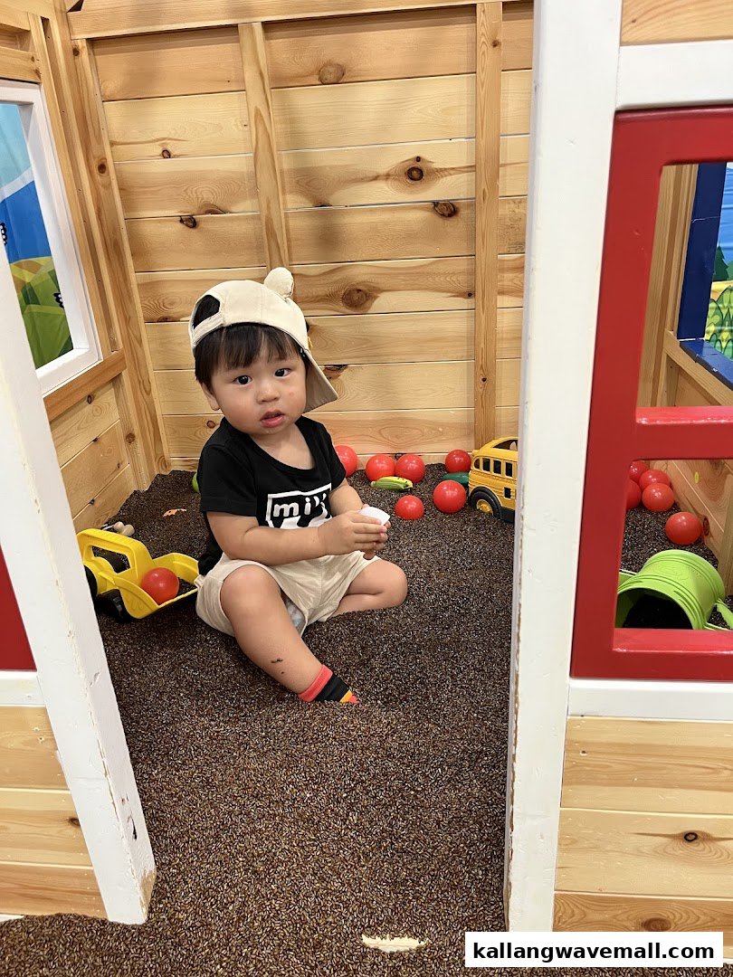 A young child in a hat plays in a wooden playhouse filled with brown pellets and toys.