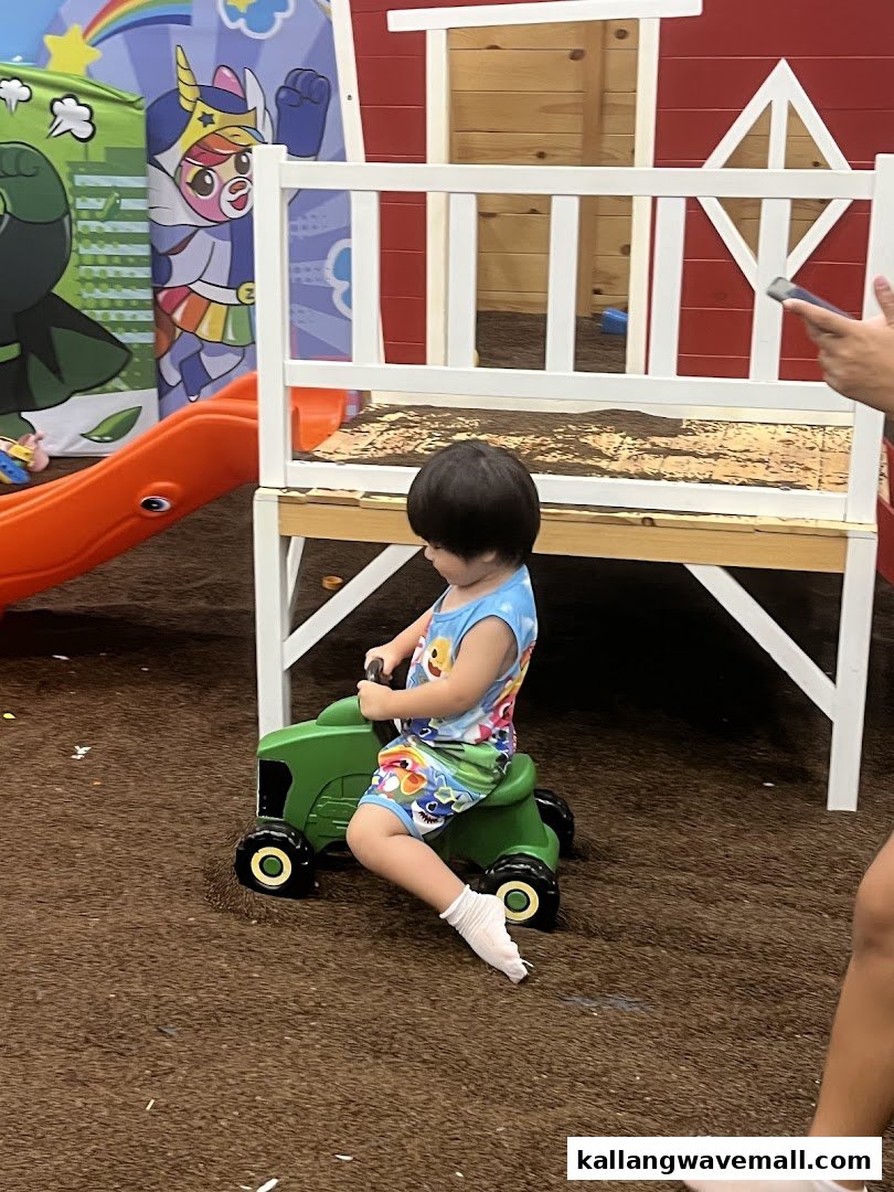 A child rides a green toy tractor in an indoor play area with a barn-themed structure and murals.