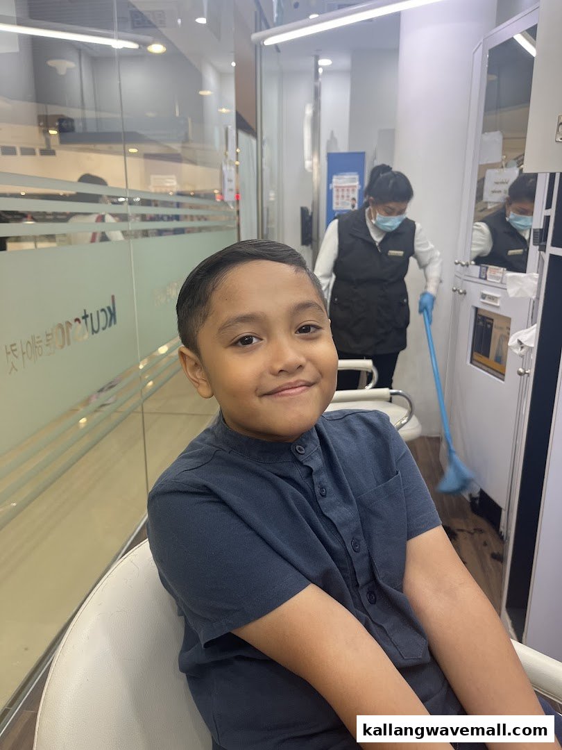 A young boy smiles after a fresh haircut at kcuts, with a staff member cleaning in the background.