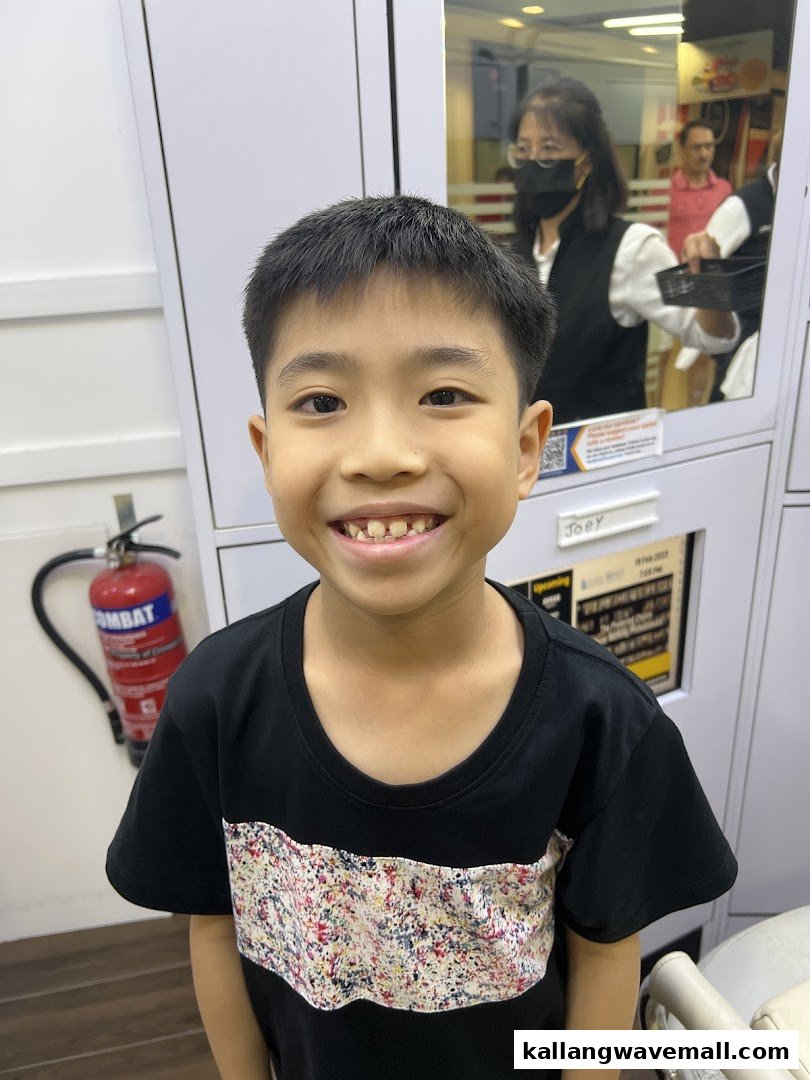 A smiling young boy inside a kcuts salon, with staff visible in the reflection.