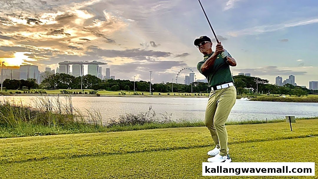 A golfer swings on a scenic course with a city skyline and lake at sunset.