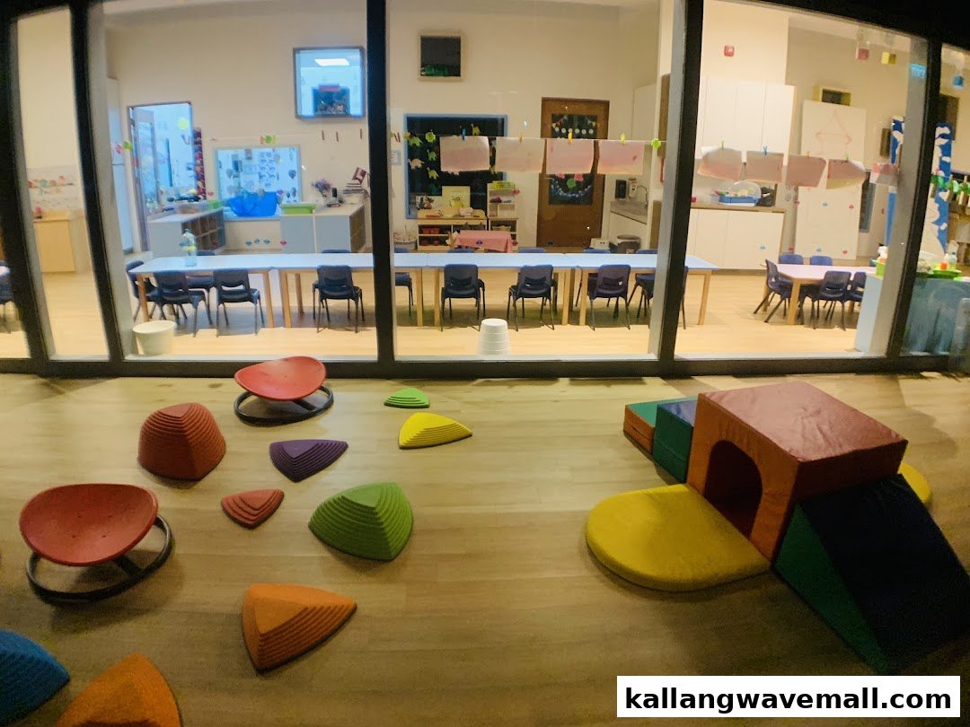 A brightly lit preschool classroom with colorful indoor play equipment visible through large windows.