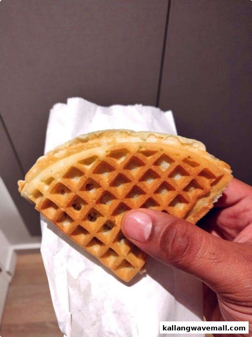 A hand holds a golden-brown, freshly baked waffle in a white paper wrapper.
