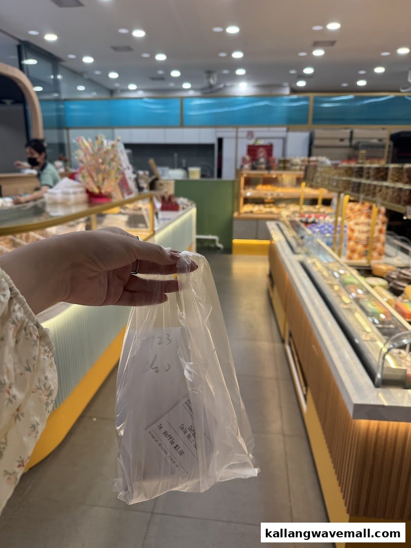 A customer holds a plastic bag with a waffle receipt inside, showcasing the interior of a bustling bakery.