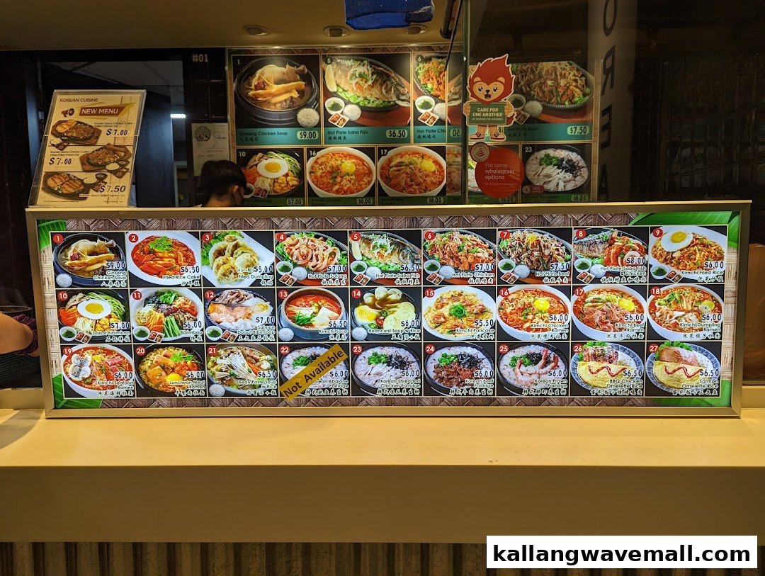 A brightly lit food display counter showcasing a variety of Korean dishes with prices at My Kampung.