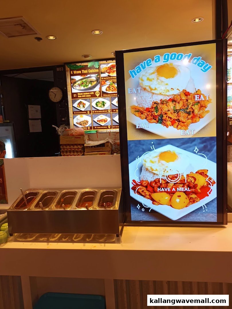 A food stall counter within 'My Kampung', featuring condiment trays and a digital display showcasing Thai dishes.