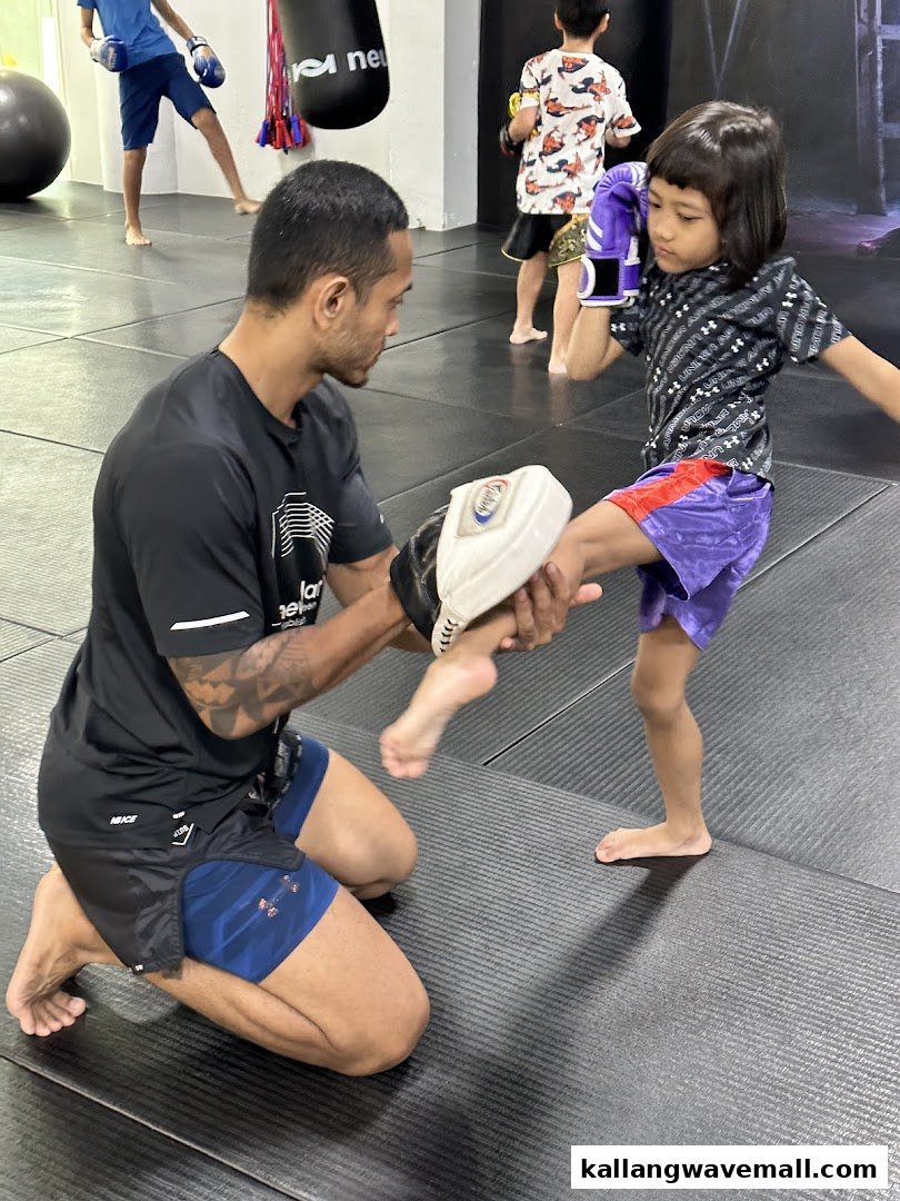An instructor guides a child practicing a kick in a martial arts gym, with other children training in the background.