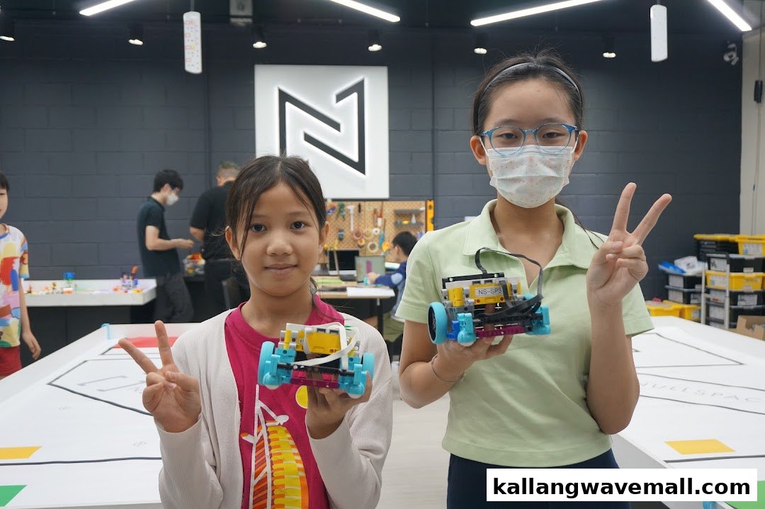 Two girls proudly display their completed robots, making peace signs inside a robotics workshop.