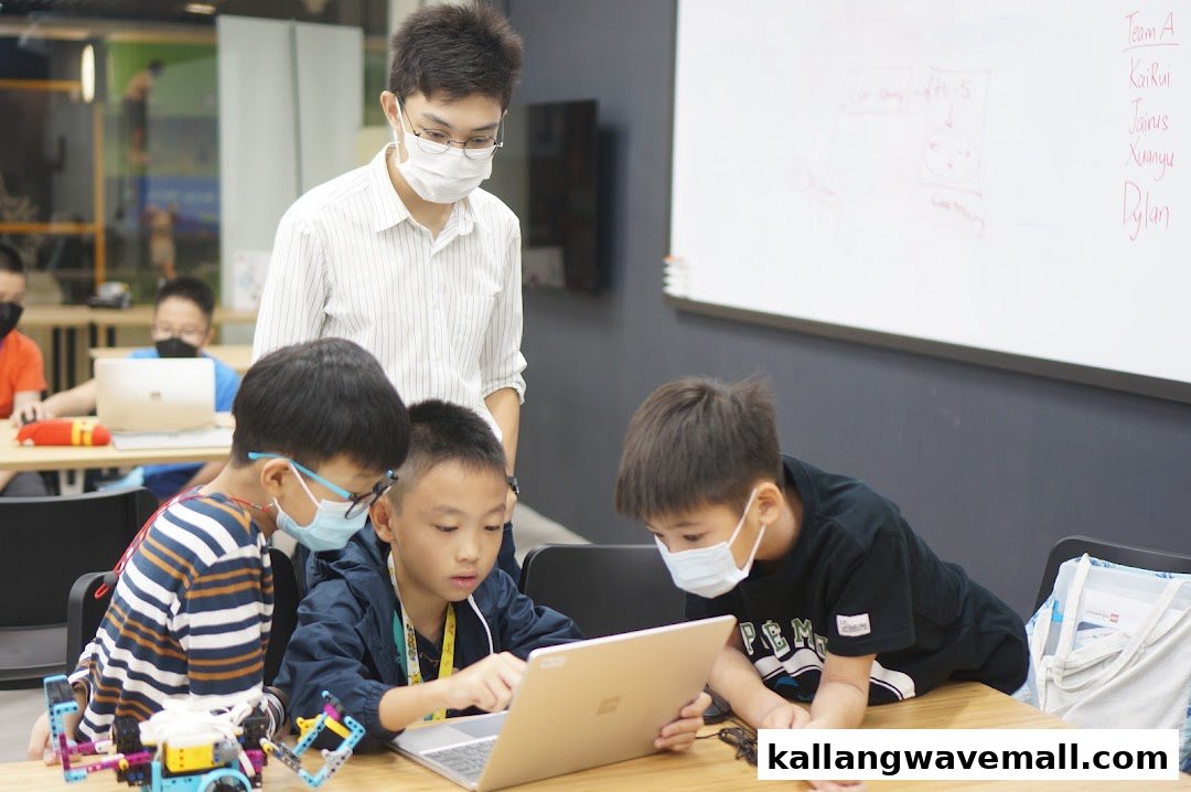 A teacher guides young students working on laptops and robotics projects in a bright classroom setting.