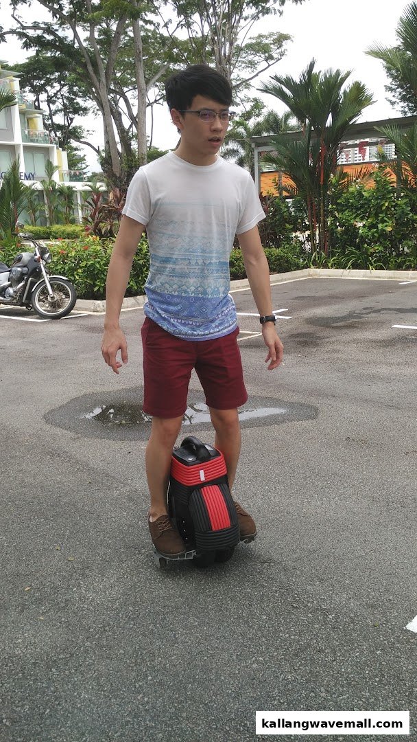 A person is riding a black and red electric unicycle on an asphalt surface outdoors.