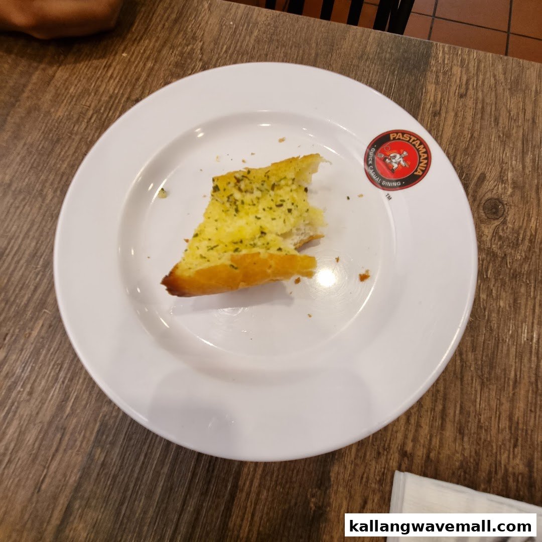 A partially eaten slice of garlic bread on a white plate with the PastaMania logo.