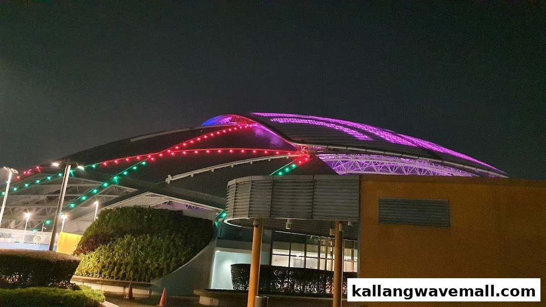 A large, modern stadium or arena illuminated with vibrant purple, red, and green lights at night.