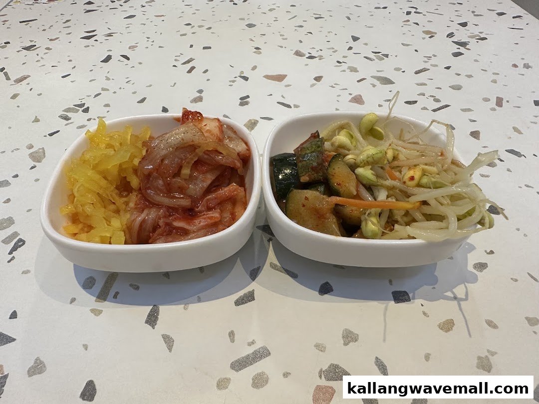 Two white bowls filled with Korean banchan, including kimchi and bean sprouts, on a speckled table.