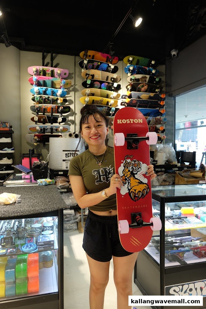 A smiling woman holds a red skateboard with a tiger graphic inside a vibrant skate shop.