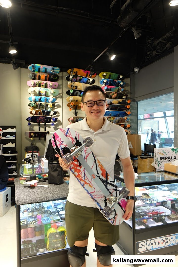 A smiling man holds a vibrant skateboard inside Skatebruh Skate Shop, surrounded by a colorful display of skateboards.
