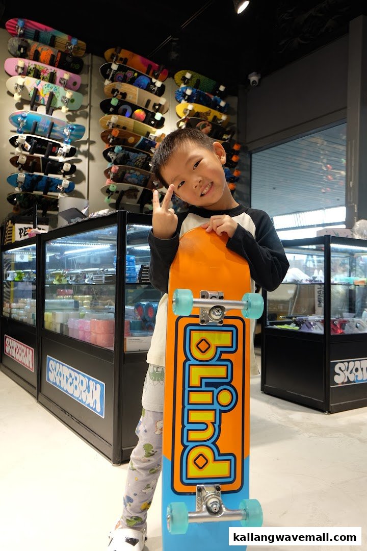 A young boy smiles, holding a skateboard inside a vibrant skate shop with many boards displayed.