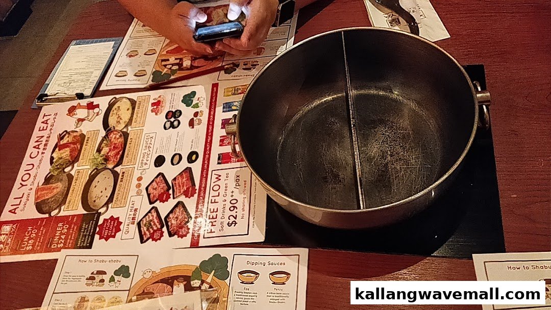 A table setting at Suki-Ya with an empty hot pot, menus, and a person using a phone.