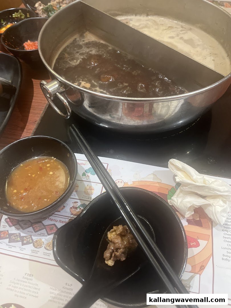 A bubbling shabu-shabu hot pot with two broths, dipping sauce, and a partial menu on a table.