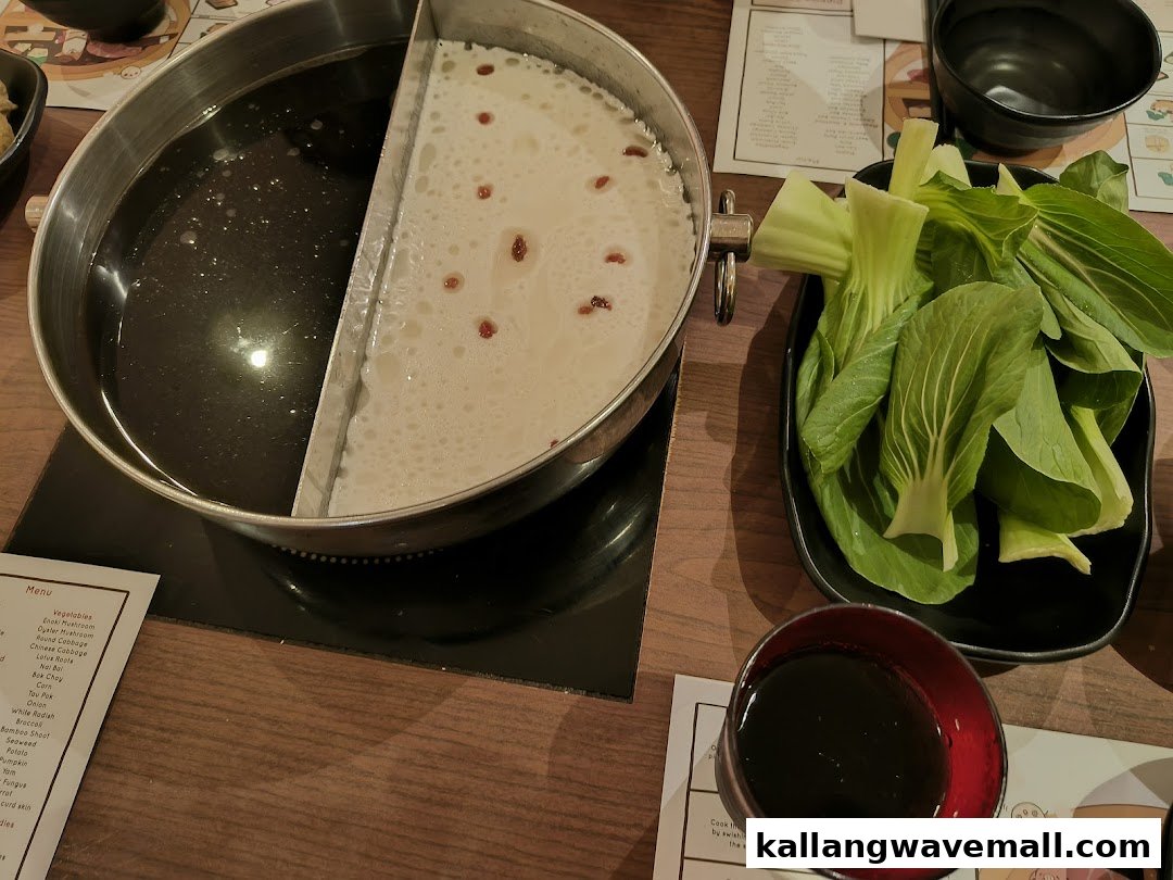 A hot pot with two simmering broths, fresh bok choy, and a dipping sauce on a wooden table.