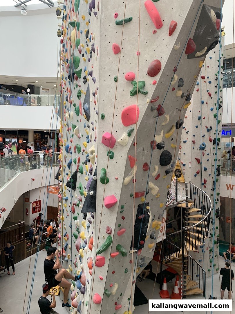 An expansive indoor climbing wall with climbers actively engaged, set within a vibrant shopping mall environment.