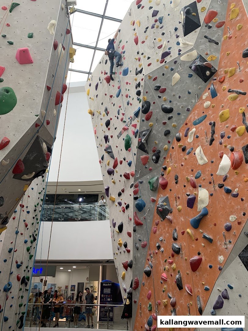 An indoor climbing gym with a person scaling a tall rock wall, surrounded by other climbing structures and observers.