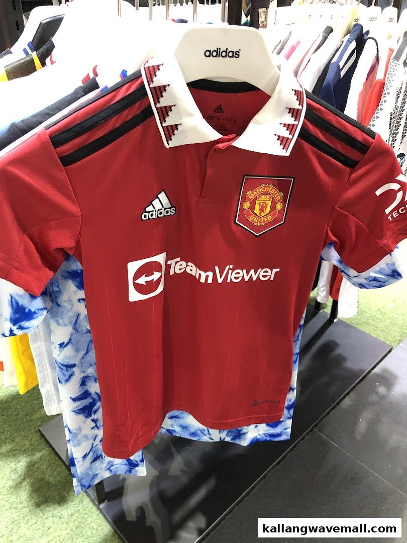 A red Manchester United football jersey with Adidas branding displayed on a hanger in a retail store.