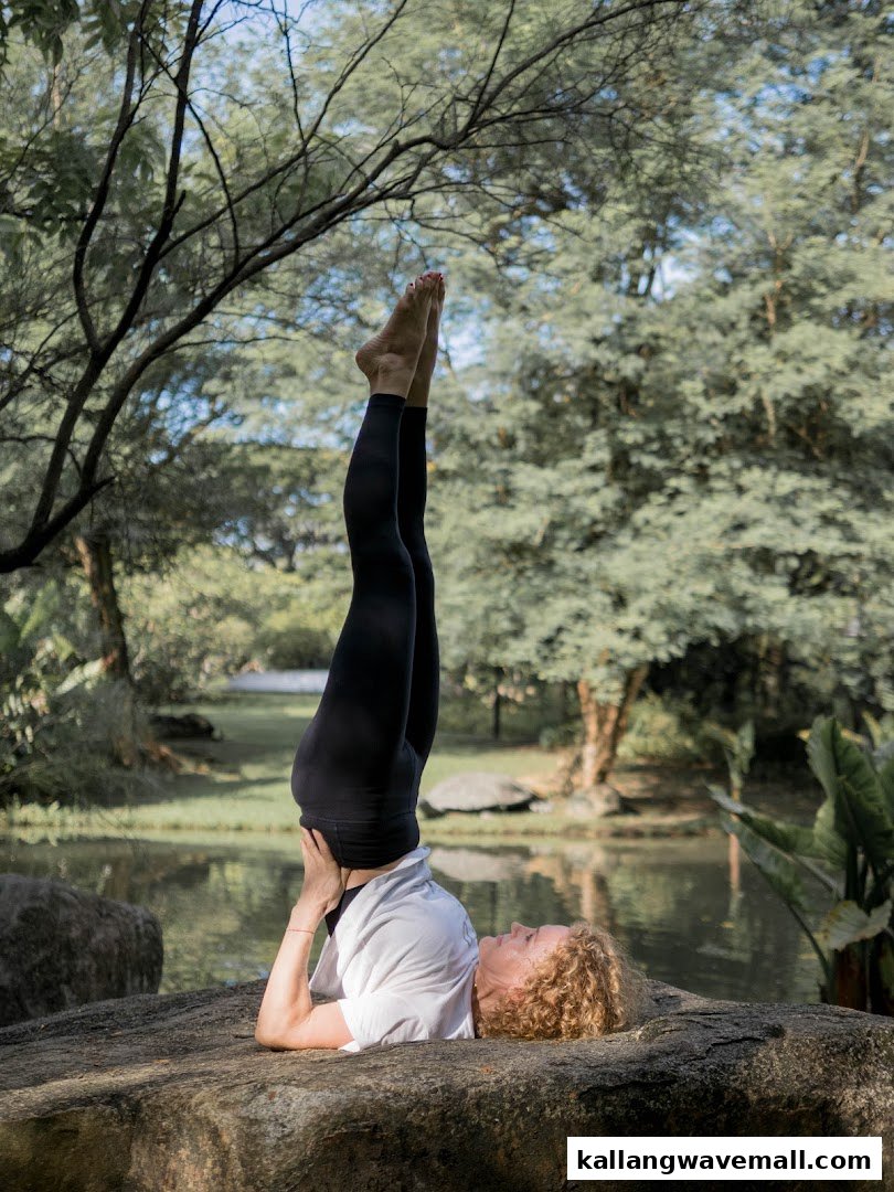 A woman performs a yoga inversion pose outdoors by a pond, surrounded by lush greenery.