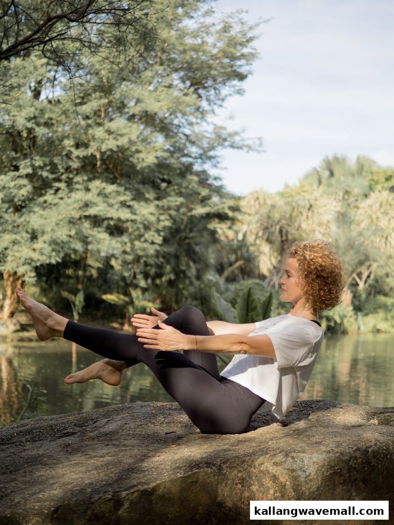 A woman performs a yoga pose on a rock by a lake, surrounded by lush outdoor greenery.