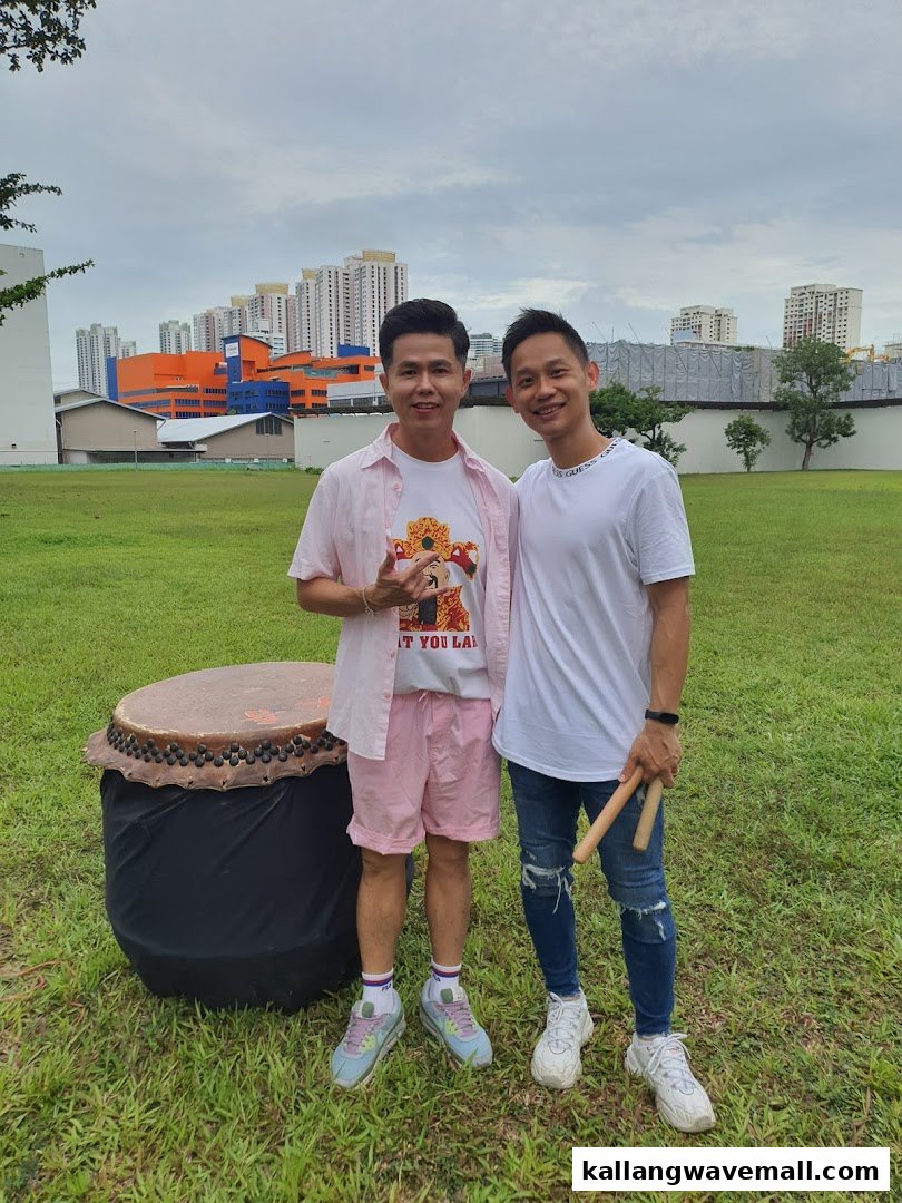 Two men pose with a large drum in a grassy field with city buildings in the background.