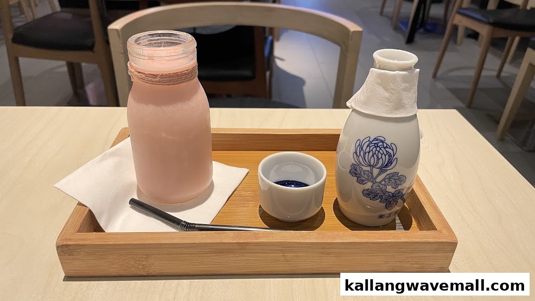 A wooden tray holds a pink drink bottle, a ceramic sake bottle, and a small cup on a table.