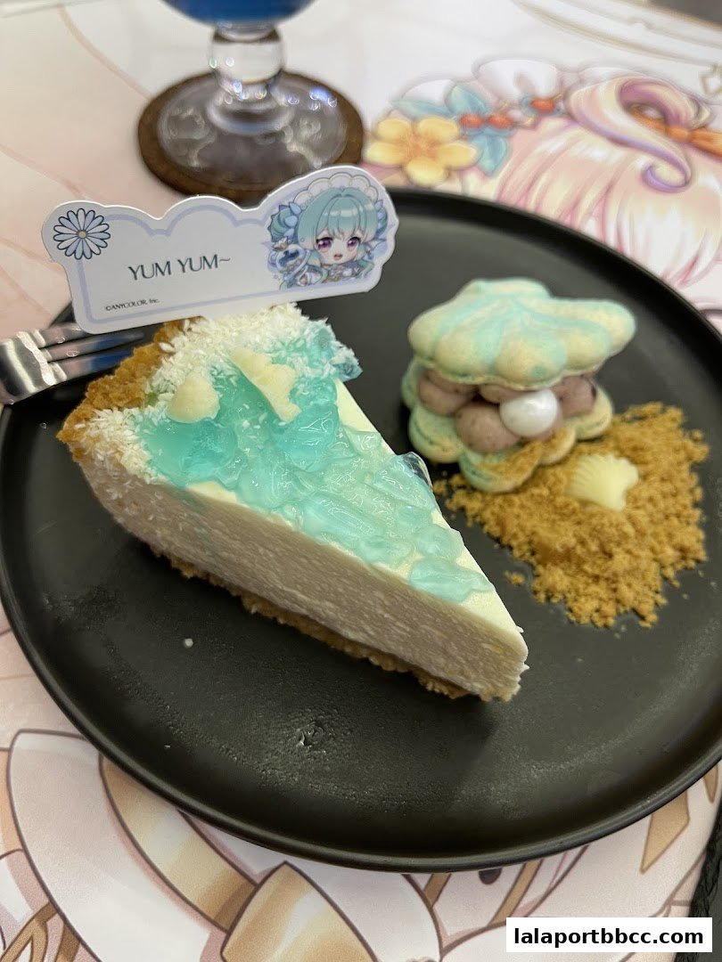 A slice of cheesecake with blue jelly and a seashell-shaped macaron on a black plate.