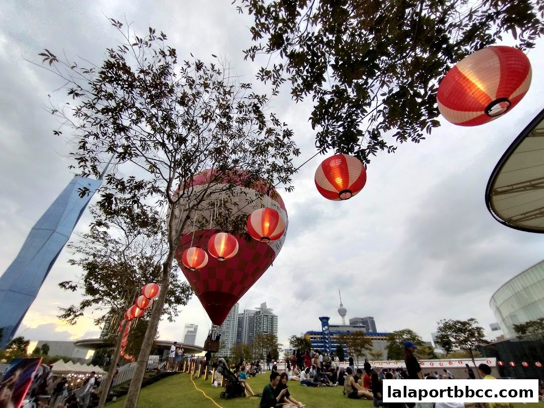 A festive outdoor scene with hot air balloons and lanterns, people relaxing on grass, and city skyline.