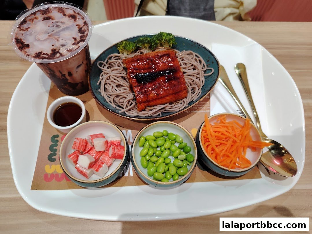 A delicious meal featuring grilled eel, soba noodles, edamame, crab sticks, shredded carrots, and a chocolate drink.